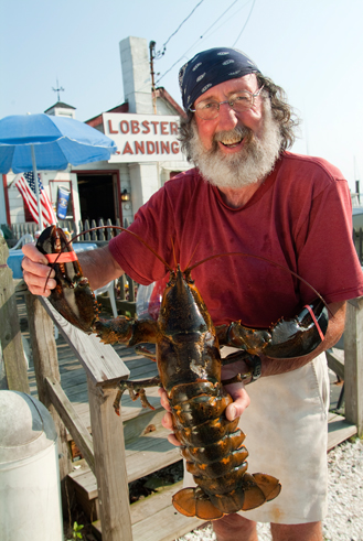 man with beard and bandana holding large lobster in front of seafood shack